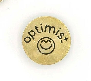 A circular brass pin with "optimist" written in black with a black smiley face.