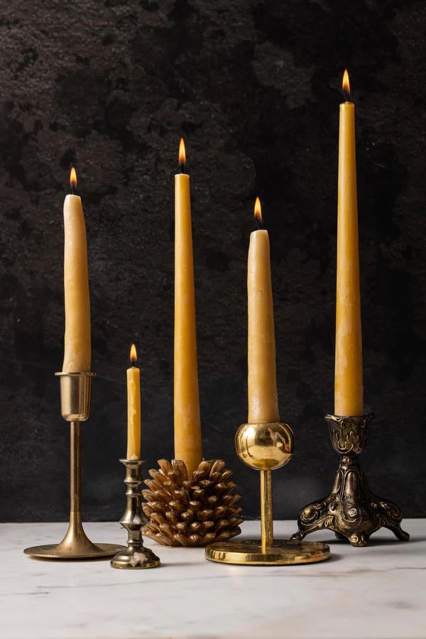 Five lit tapered beeswax candles are on display at varying heights against a black background. Each is placed in a different style candleholder.