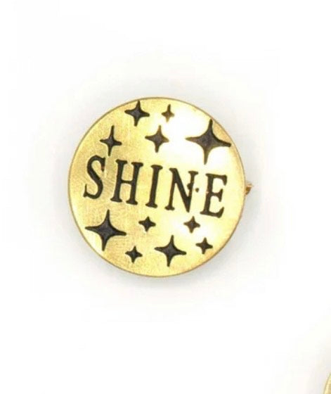 A circular brass pin with black stars and "SHINE" written in black.