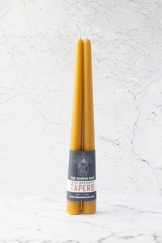 Two tapered yellow candles with white wicks are wrapped with grey paper that reads "100% Beeswax Tapers". 